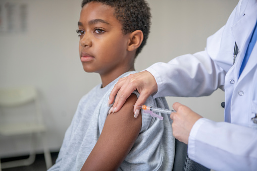 A young boy of African decent sits up on a exam table with a gown on as his female doctor administers his vaccine.  He has his sleeve rolled up and a neutral expression on his face as she injects the needle.