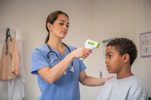 A female doctor of African decent holds up a thermometer gun in front of her young male patients head as she checks his temperature.  She is wearing blue scrubs and the boy is sitting up on the exam table in a gown and a neutral expression on his face as he lets her test him.