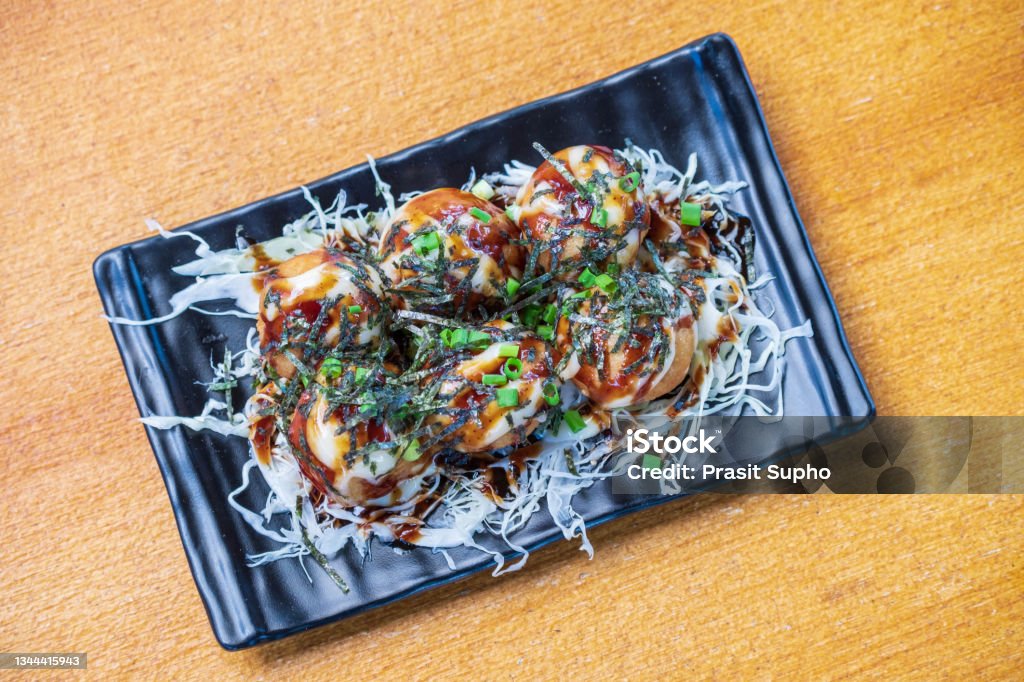Octopus Fillet Takoyaki Garnish with seaweed. chopped white cabbage and scallions in a black dish, Saimoden, circled on a wooden table Photography Stock Photo