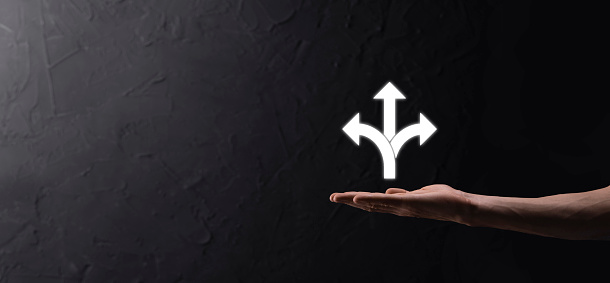 Male hand holding icon with three directions icon on dark background. Doubt, having to choose between three different choices indicated by arrows pointing in opposite direction concept. Ways.