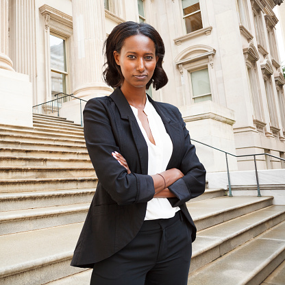 A well dressed woman with arms folded standing in front of a courthouse or municipal bulding looks at viewer. Could be a lawyer, business person etc.