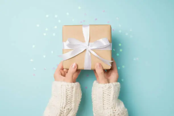Photo of First person top view photo of female hands in white pullover giving craft paper giftbox with white ribbon bow over shiny snowflakes confetti on isolated pastel blue background