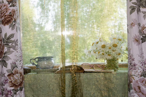 On the windowsill behind a transparent green curtain is a bouquet of daisies, an open book and glasses, and a cup of coffee.