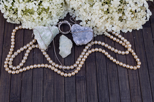 On a wooden background lies three semiprecious stones of aquamarine, prenite, celestine. Nearby are beads, a silver ring and a chain and white hydrangea flowers.