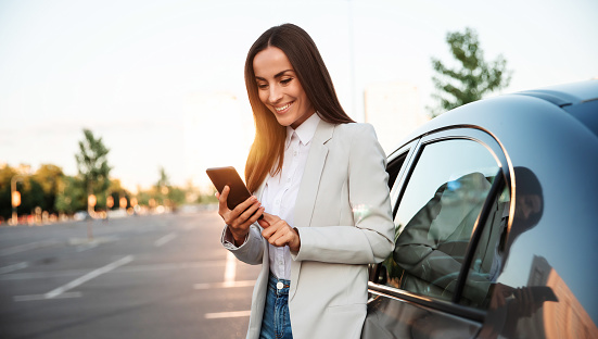 Una mujer atractiva y sonriente exitosa con ropa inteligente formal está usando su teléfono inteligente mientras está de pie cerca de un automóvil moderno al aire libre. photo