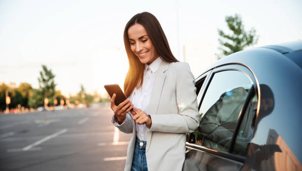 una mujer atractiva y sonriente exitosa con ropa inteligente formal está usando su teléfono inteligente mientras está de pie cerca de un automóvil moderno al aire libre. - car fotografías e imágenes de stock