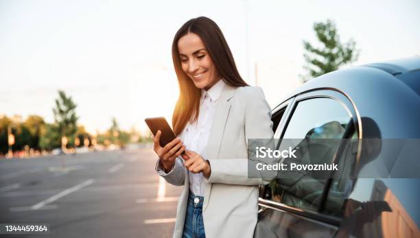 Successful Smiling Attractive Woman In Formal Smart Wear Is Using Her Smart Phone While Standing Near Modern Car Outdoors Stock Photo - Download Image Now