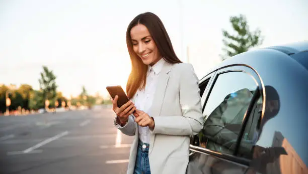 Photo of Successful smiling attractive woman in formal smart wear is using her smart phone while standing near modern car outdoors
