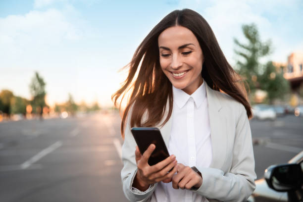 istock Successful smiling attractive woman in formal smart wear is using her smart phone while standing near modern car outdoors 1344459374