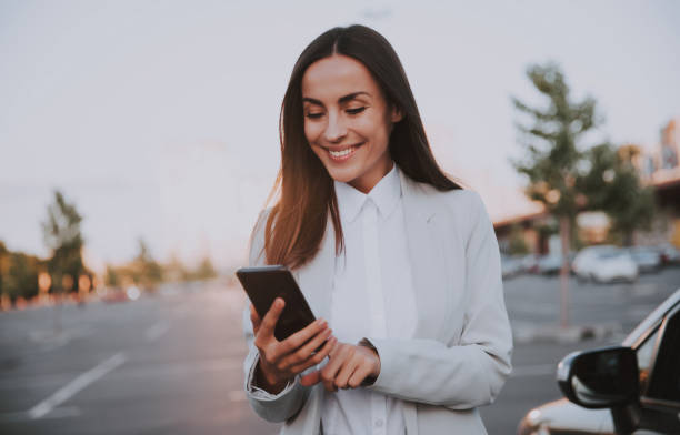 istock Successful smiling attractive woman in formal smart wear is using her smart phone while standing near modern car outdoors 1344459519