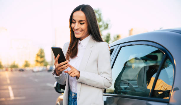 Successful smiling attractive woman in formal smart wear is using her smart phone while standing near modern car outdoors stock photo