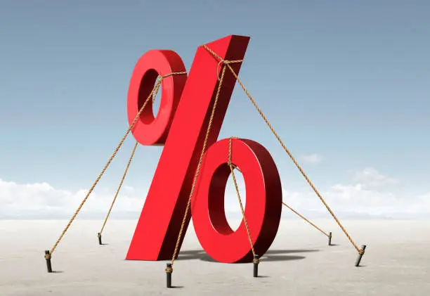 A tall red percent sign is restrained by ropes attached to stakes in the ground conveying concept of artificially low rates restrained by Federal Reserve policies.