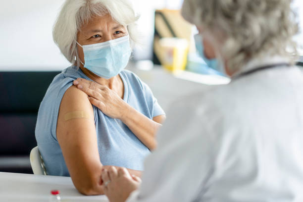 Senior Patient Successfully Receiving Her Vaccination A senior patient of Filipino decent holds up her sleeve and smiles as she looks up at her doctor after receiving her vaccination.  She is dressed casually in a blue sweater, has a bandage on her arm and is wearing a medical masks to protect her from the virus. flu vaccine photos stock pictures, royalty-free photos & images