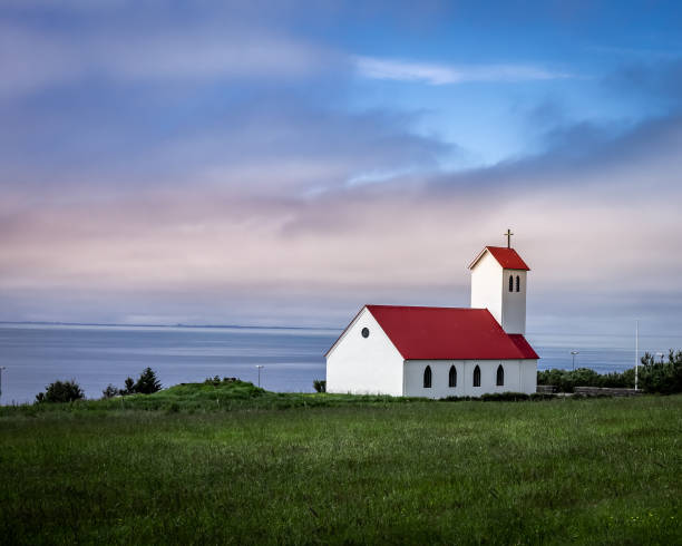 Hafnarfjordur, Iceland - July 17, 2021:\nA scenic Icelandic landscape. Traditional church with red rooftop, situated on a green hill, pink clouds in the sky.