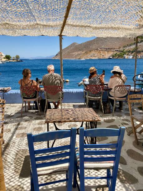 Seaside cafe under the net which is making beautiful pattern of shades on the stone floor. Unrecognizable people are sitting by the table towards the Aegean sea. Cozy holiday morning