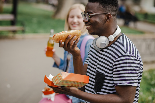 Young African-American man and Caucasian woman are eating a hot dog in the park and having a great time