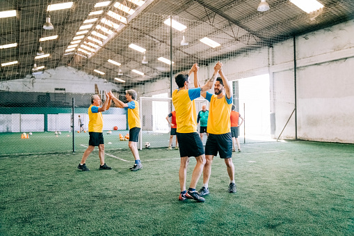 Smiling football players giving each other high fives on indoor field after scoring a goal. Successful soccer players celebrating after winning the match.