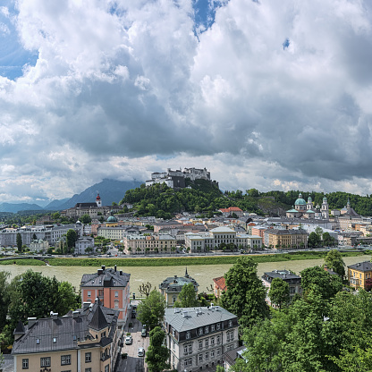 Salzburg, Austria - May 25, 2017: View over the Salzach river, Old Town and Hohensalzburg fortress with low cumulus clouds above them. View from the fortified wall at the Kapuzinerberg mountain.