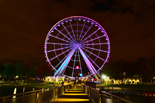 Long exposure of Montreal Grand Ferris Wheel in Old Port at the twilight blue hour. Background is cloudy moody sky. Foreground is fall color trees and a bridge.