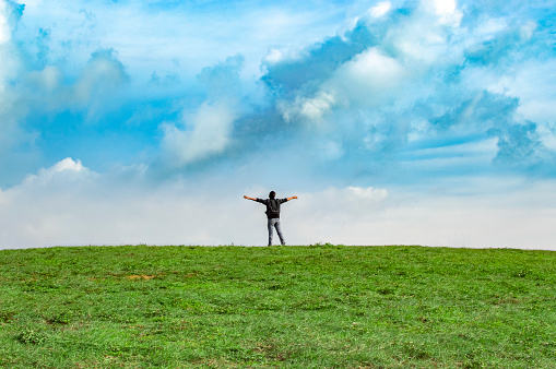 A backpacker on a hill with blue sky and copy space, man backpacking on a green hill with copy space, concept of successful man spreading his hands