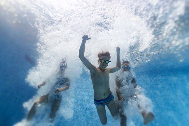 Teenage boys and a teenage girl are having fun swimming underwater in pool. The kids are jumping into the pool and they are smiling at the cameraShot with Canon R5.