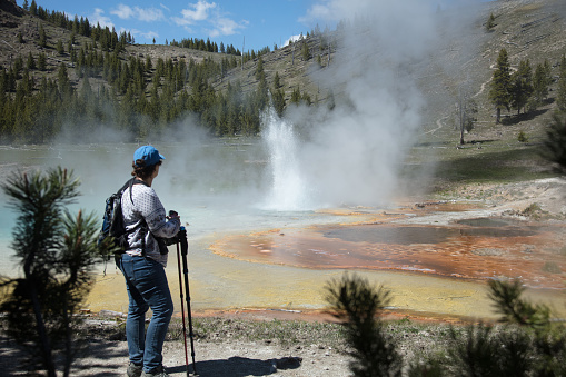 Yellowstone National Park - Hike to Imperial Geyser. Woman standing in front of geyser looking at it erupting. Shot from behind woman. Yellow, gold, orange, and turquoise colors.