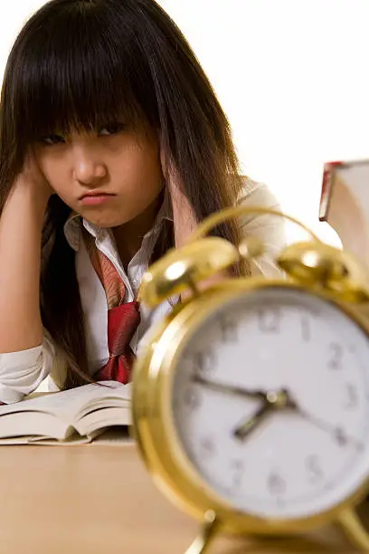 Young Chinese girl wearing school uniform sitting in front of thick textbooks with hands on sides of head with a pouting face expression with an alarm clock in front