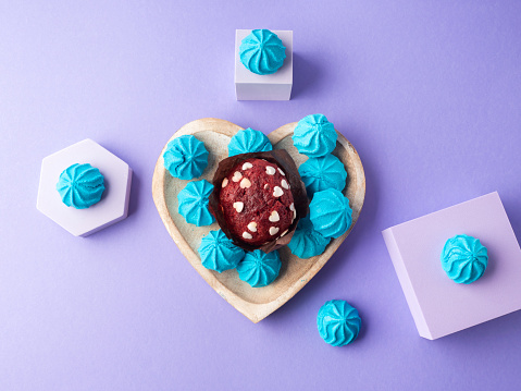 Heart shaped wooden dish with blue sweet meringues and red velvet muffin on purple background. Flat lay with geometric shapes podiums