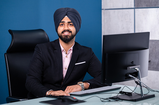 Indoor image of Asian/ Indian Young Sikh businessman in formal dress with turban working on computer and using smartphone at office.
