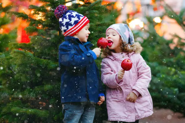 Photo of Two little smiling kids, preschool boy and girl eat sweet sugared apple on German Christmas market. Happy siblings children with lights on background and xmas trees. Family funny brother and sister.