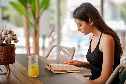 Young pretty woman is sitting and drink a glass lemonade while she is reading a book . High quality photo