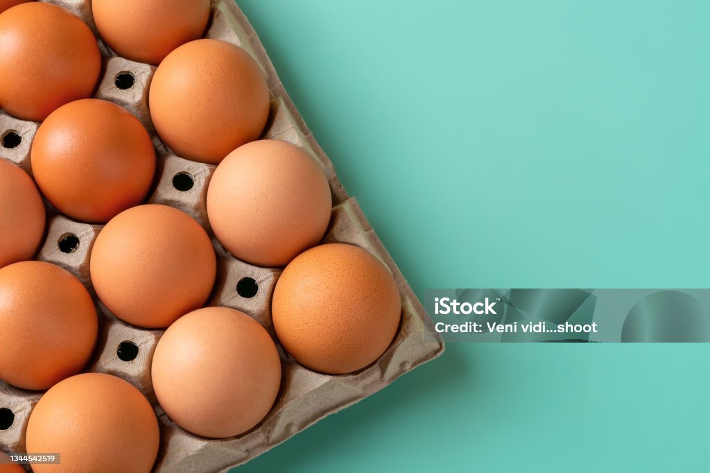 Tray with brown chicken eggs against turquoise background. Copy space. Copy space at cardboard tray with  brown chicken eggs against turquoise background. Raw organic hen eggs with brown shell as protein ingredient for healthy eating and breakfast. Farm eggs for Easter. Top view. Packaging Stock Photo