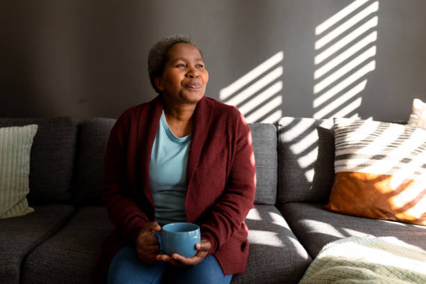 entspannte afroamerikanische seniorin sitzt auf dem sofa und trinkt kaffee - women mature adult black american culture stock-fotos und bilder