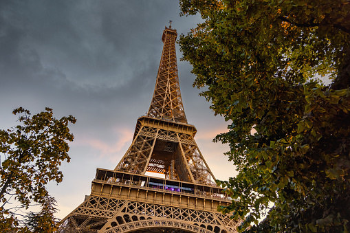 Eiffel Tower in Paris under moody dramatic sunset twilight skyscape between green summer trees. Eiffel Tower, Champ de Mars, 7th Arrondissement, Paris, France, Europe