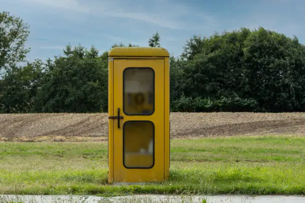 Single old yellow phone booth in retro style in an abandoned place with grass, overgrown with weeds and wildflowers on a summer sunny day.