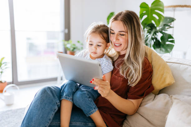 Mother and daughter using a digital tablet together Mother and daughter using a digital tablet together. They are sitting on the sofa at home. The daughter is sitting on his mothers lap. Very hapy and smiling. Close up with Tight crop one parent stock pictures, royalty-free photos & images