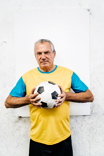 Senior man holding soccer ball against white wall. Active senior male player in sportswear standing at indoors soccer field.
