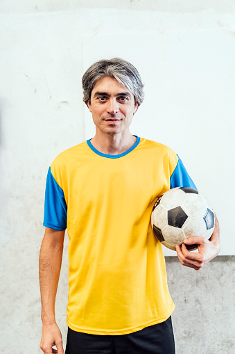 Portrait of a confident mature male soccer player holding a ball against white wall. Soccer player inside a soccer court with ball.