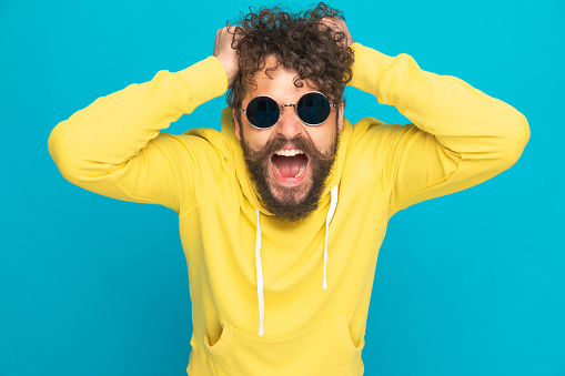 angry young man with glasses and yellow hoodie ripping out hair and screaming while standing on blue background in studio