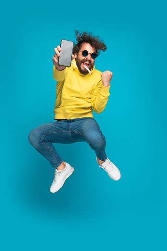 excited young man with long beard and moustache showing black empty screen, holding fist up and celebrating, jumping and screaming on blue background in studio