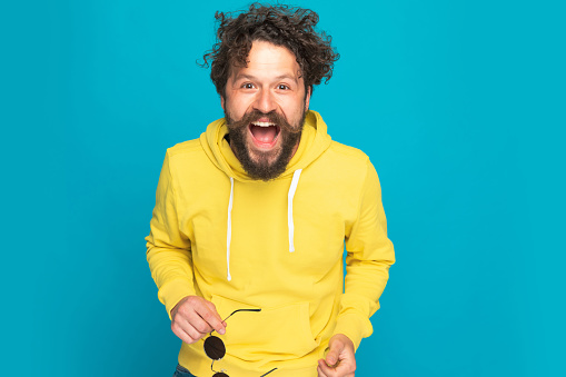 excited unshaved casual guy having fun, smiling and laughing while standing and posing on blue background in studio