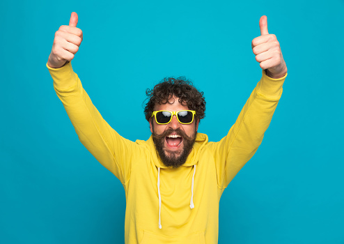 excited young casual man in yellow hoodie with sunglasses making thumbs up gesture and laughing on blue background in studio