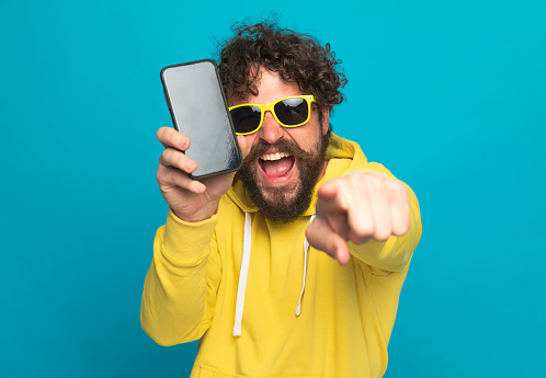 excited young man with curly hair wearing yellow hoodie and sunglasses laughing, pointing finger and showing blak empty screen while posing on blue background in studio