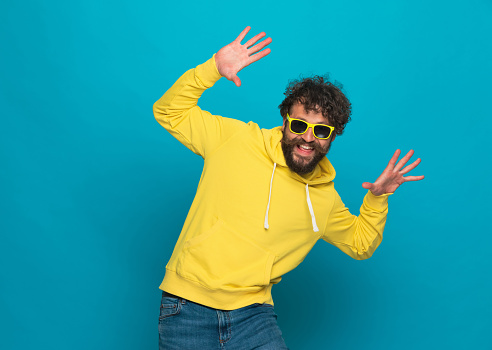 unshaved funny man with moustache and beard holding arms in the air, dancing, laughing and having fun against blue background in studio
