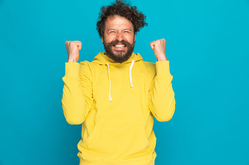 happy casual man in yellow hoodie holding fists in the air and celebrating victory on blue background in studio