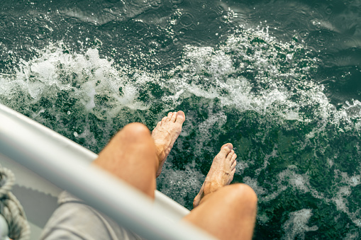 Personal perspective of a sailing man, sitting on his sailboat own a sunny day. Point of view of his legs and bare feet by the water.