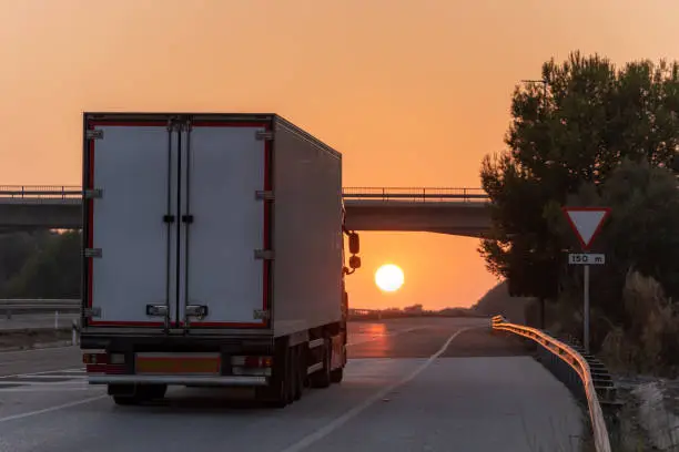 Photo of Truck with refrigerated semi-trailer driving on the highway with the sun in front of it at sunset.