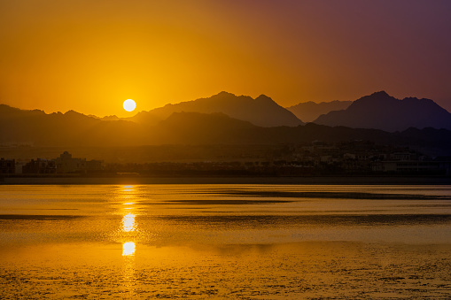 Scenic, picturesque orange sunset over black desert mountains of Egypt with reflection of sunlight in water of red sea with coastline and colorful sky in summer or autumn vacation season. Horizontal