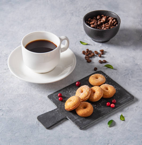 delicious breakfast with freshly brewed coffee and mini donuts in powdered sugar and lingonberry berries on a black slate on a blue background - coffee cup bean sugar imagens e fotografias de stock