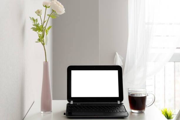 istock Modern workspace with blank screen laptop, frame, coffee cup and vase on white table against the background of a window and a light wall 1344626667